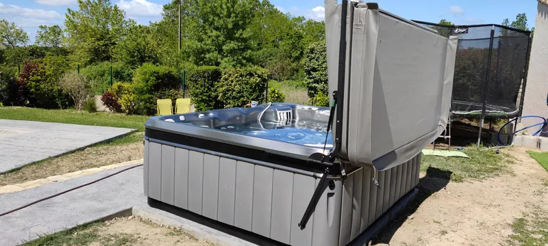 Spa gris sur sa dalle béton, couvercle relevé, trampoline derrière