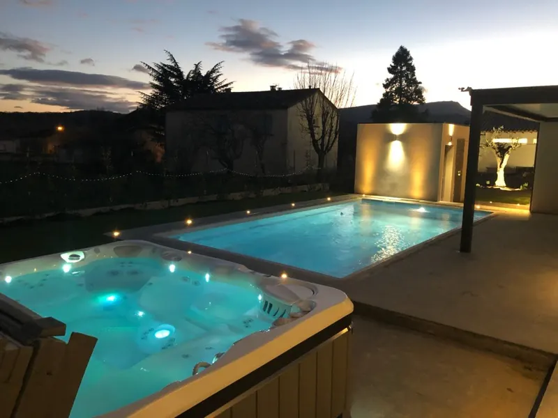 Spa blanc et piscine bleue éclairés, sur une terrasse sous pergola noire