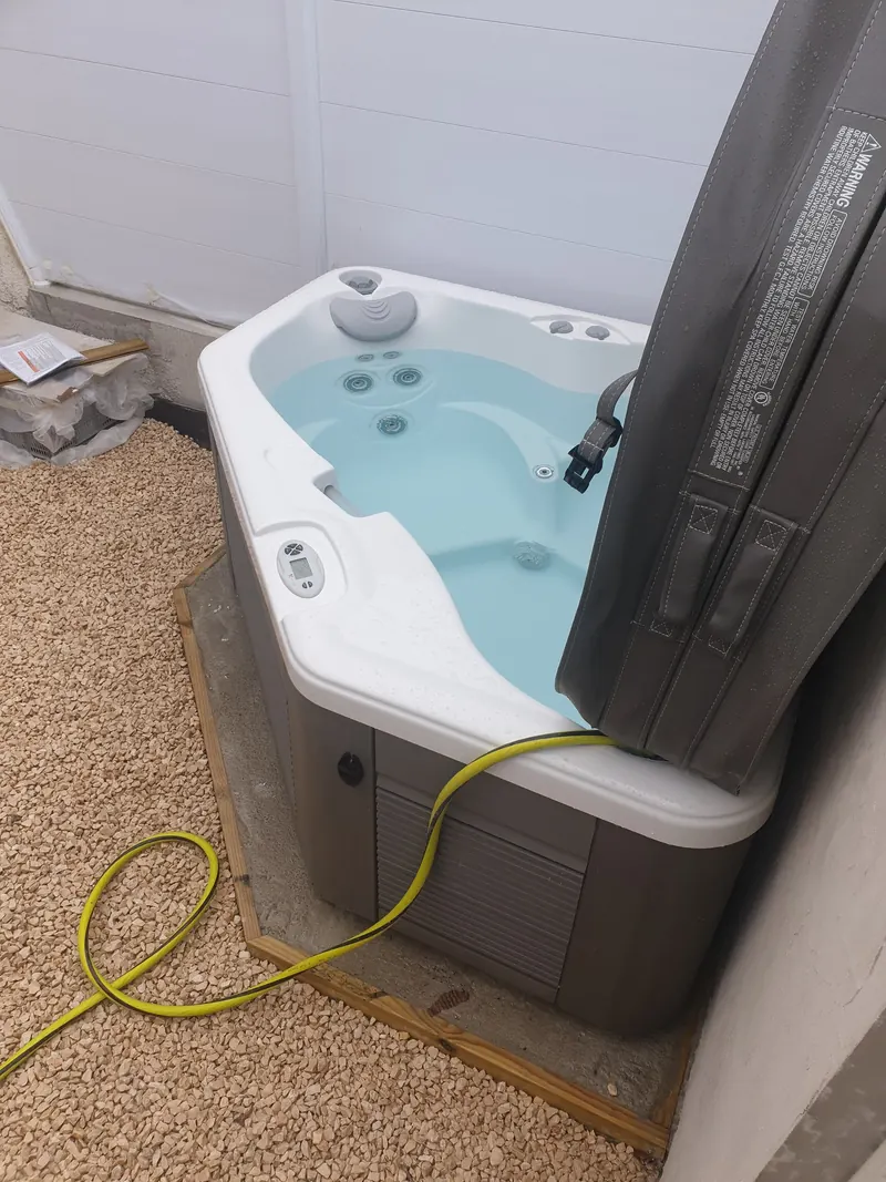 Spa d'angle blanc et gris en eau, couvercle relevé, tuyau d'arrosage jaune posé sur le bord, au Grau-du-Roi