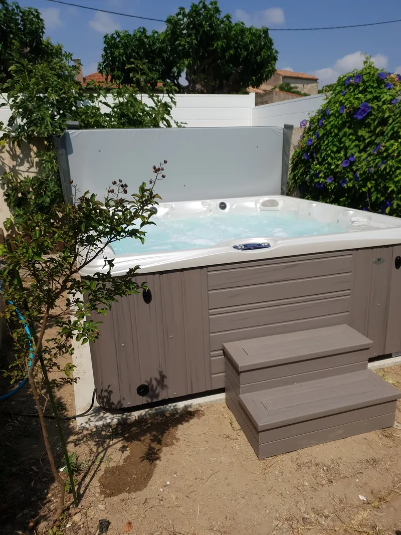Spa à habillage gris rempli, couverture relevée, contre une clôture blanche dans un jardin à Loupian