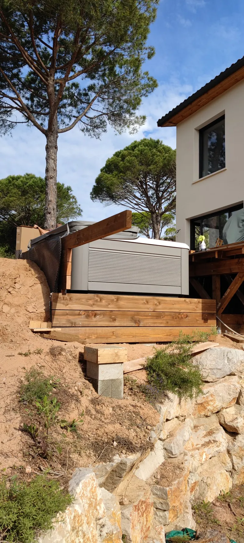 Spa sous son couvercle gris, posé sur des planches au bord de la pente, maison à droite