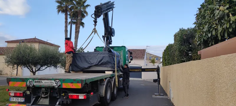 Spa sous bâche noire sur le plateau d'un camion-grue, deux personnes autour des sangles