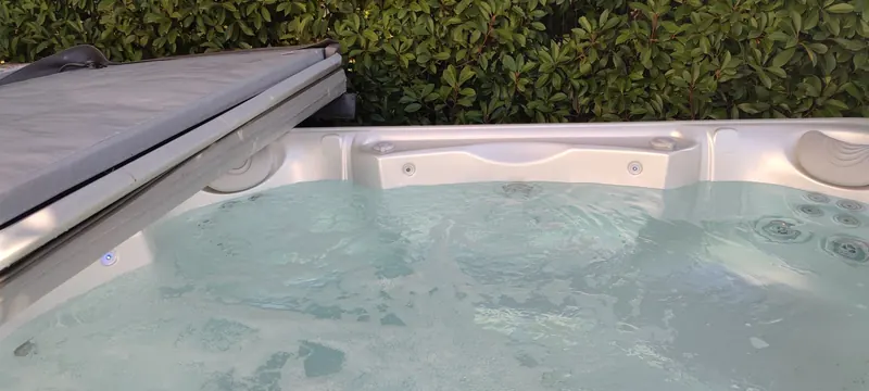 Spa blanc à l'eau turquoise, bâche entrouverte, haie verte tout autour