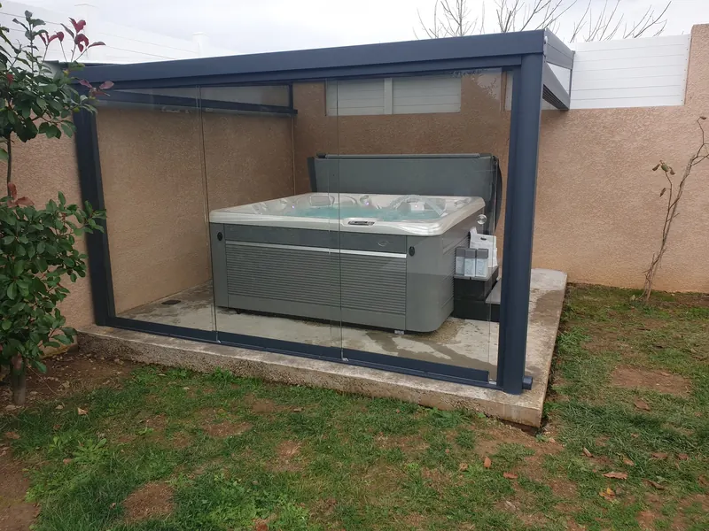 Pergola aux parois vitrées abritant un spa gris sur dalle béton au fond d'un jardin à Thézan-lès-Béziers