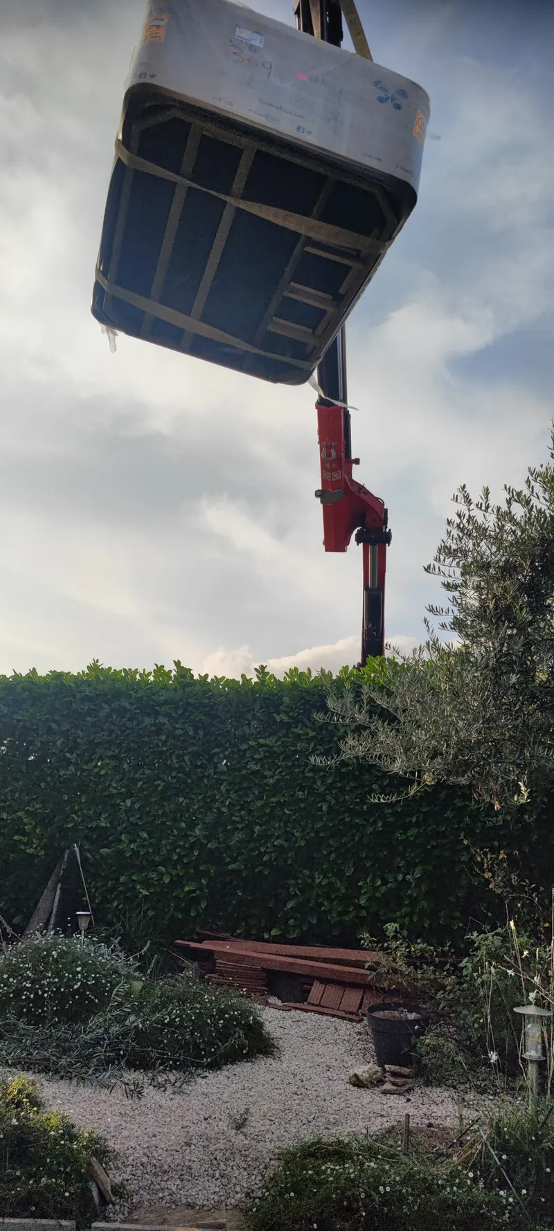 Spa ovale gris suspendu à la grue rouge, vu de profil au-dessus du jardin et de sa haie vive