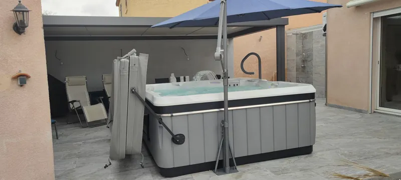 Spa blanc et noir sur une terrasse couverte, pergola grise et parasol bleu au-dessus